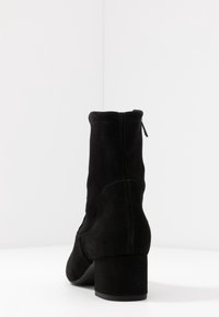 Stivale alla caviglia in suede nero con tacco a blocco e zip posteriore, fotografato su uno sfondo bianco con angoli.