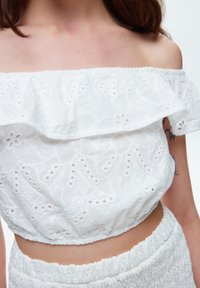 Weißes Off-Shoulder-Oberteil mit Broderie Anglaise-Details, gerafftem Ausschnitt und elastischem Saum. Weicher, strukturierter Stoff mit floralem Muster.