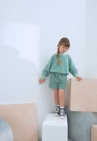 Ensemble de sweat-shirt rayé vert et de short avec une taille élastique, associé à des baskets montantes noires et blanches, placé sur des blocs blancs et pastel.