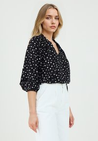 Blusa nera con pois bianchi, scollo a V e maniche arricciate, abbinata a pantaloni bianchi a vita alta. Tessuto leggero e vestibilità comoda.