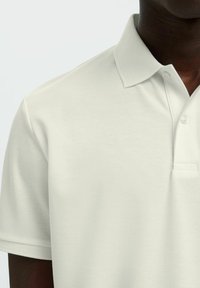 Homme portant un polo à manches courtes couleur crème avec un col texturé et une patte de boutonnage, visible du torse au cou.
