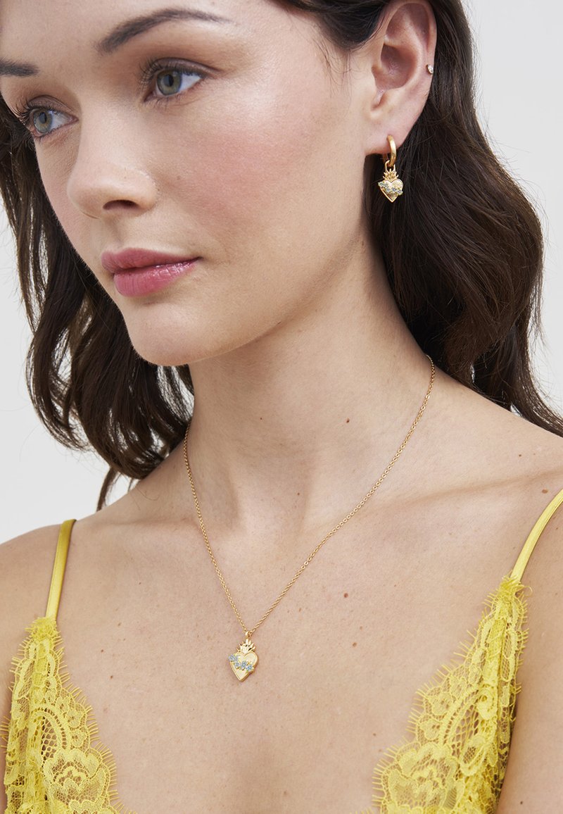 Žena s tamnom kosom, koja nosi žuti čipkasti top, zlatni ogrlicu s privjeskom u obliku srca s cvjetnim dizajnom i odgovarajuće zlatne naušnice u obliku srca.