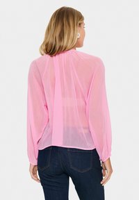Blouse légère et transparente de couleur rose pâle avec des manches longues et un dos en ouverture en forme de clé. Le tissu semble délicat avec une texture lisse. Portée sur un jean noir.
