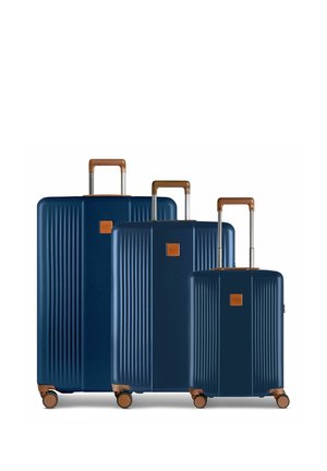 Trois valises rigides bleu marine avec des poignées et des roues marron, disposées côte à côte du plus grand au plus petit.