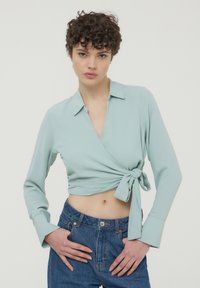 Blusa avvolgente blu chiaro con maniche lunghe, legata in vita, con colletto e texture liscia, abbinata a jeans in denim a vita alta.