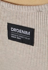 Czarna metka z białym napisem "DRDENIM Denim & Streetwear Since 2004" przyszyta do beżowej prążkowanej tkaniny.