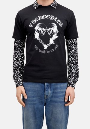 Osoba ubrana w czarny T-shirt z białymi czaszkami tworzącymi kształt serca oraz napisami „THE KOOPLES” i „Till death do us part”, połączony z długimi rękawami w wzory.