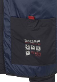 N2A New Zealand jas in donkerblauw nylon, met een voorvak met een rode accent. Toont symbolen voor temperatuurregeling, winddicht en waterafstotend.