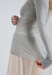 Siva majica z dolgimi rokavi, izdelana iz mehke tkanine, z zaobljenim robom in odprtinami za palce, nosena preko svetle krila.