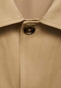 Tan button-up collar with a smooth fabric texture. Features a round, dark button and precise stitching along the edges.
