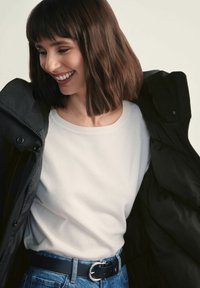 Kobieta uśmiechnięta, patrząca w dół, ubrana w biały sweter, niebieskie dżinsy z czarnym paskiem oraz czarną kurtkę zarzuconą na ramiona.