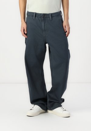 Personne portant un pantalon large bleu foncé et des baskets blanches, debout devant un fond clair uni.