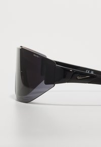 Gafas de sol con visera negra y un marco brillante, lentes oscuros con degradado, diseño anguloso y un pequeño logo de Nike en el lateral.