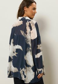 Frau, die ein lockeres, langärmliges Hemd mit abstraktem Muster in Marineblau, Beige und Weiß trägt, steht mit teilweise gedrehtem Rücken.