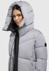 Giacca imbottita grigia chiara con un grande cappuccio strutturato, chiusura con zip nera e finitura opaca. Presenta un dettaglio a righe trapuntate orizzontali e un accento con logo.