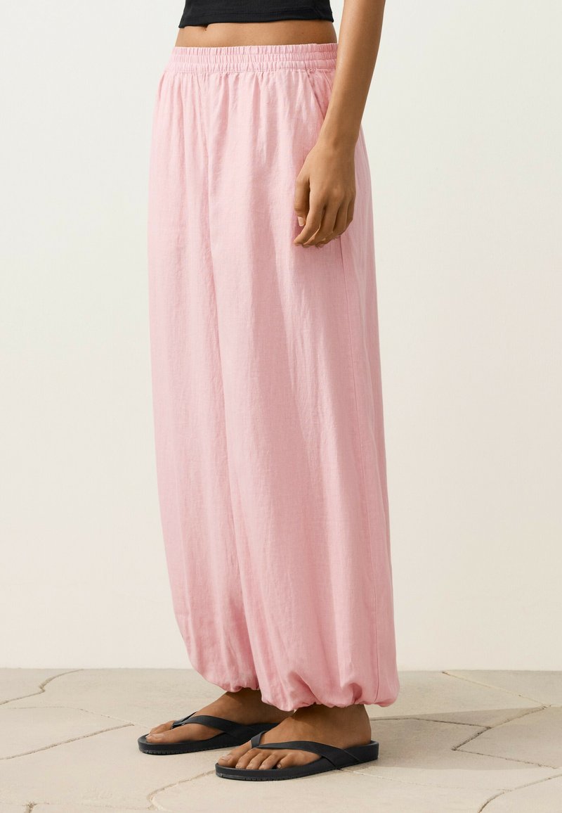 Femme portant un pantalon ample rose clair avec des poignets élastiques, des tongs noires et un top court noir, debout sur un sol carrelé.