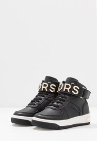 Černé vysoké tenisky vyrobené z umělé kůže, s tlustou bílou podrážkou, zlatou páskou s nápisem a černými tkaničkami.