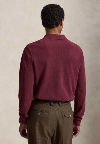 Homme portant une chemise bordeaux à manches longues rentrée dans un pantalon marron à carreaux, vu de dos sur un fond neutre.