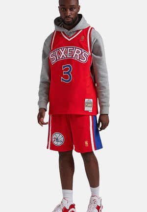 Homme portant un maillot de basket rouge des Philadelphia Sixers numéro 3, un short assorti, un sweat à capuche gris, des chaussettes blanches et des baskets rouges et blanches.