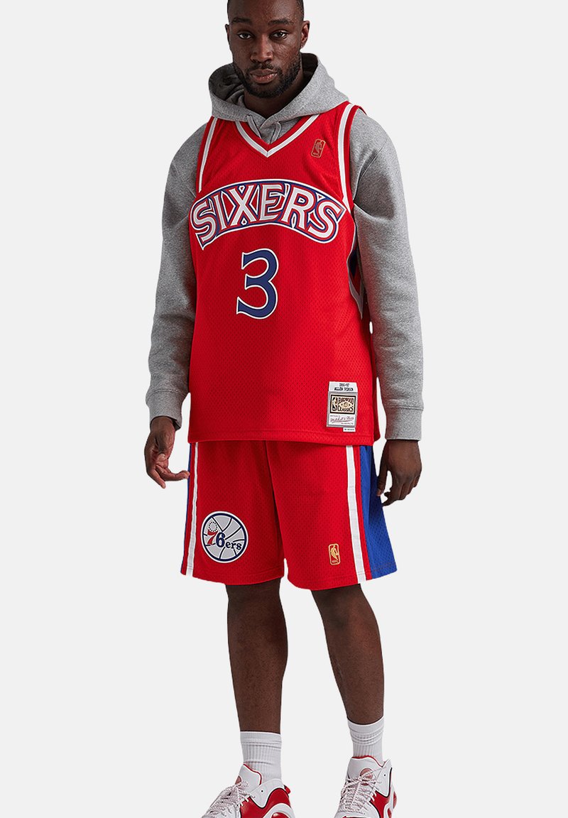 Hombre que lleva puesta una camiseta roja de baloncesto de los Philadelphia Sixers con el número 3, pantalones cortos a juego, sudadera gris con capucha, calcetines blancos y zapatillas rojas y blancas.