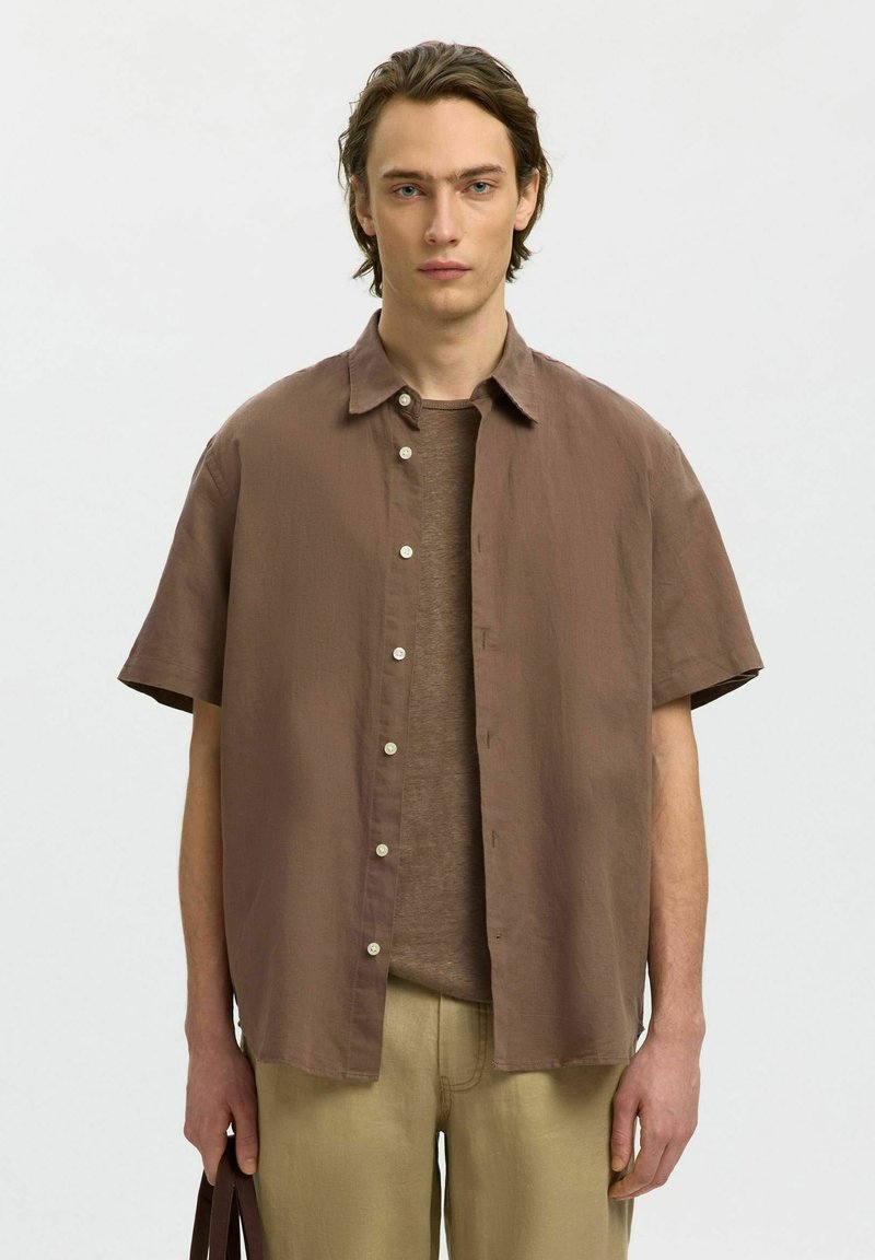 Jeune homme portant une chemise à manches courtes marron boutonnée sur un t-shirt marron, tenant un sac à poignée marron foncé, debout devant un fond uni.