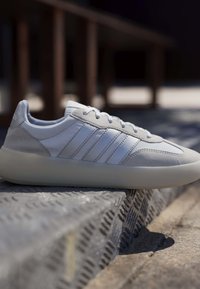 Baskets blanches avec accents en daim gris et bandes blanches reposant sur des marches métalliques texturées en extérieur sous la lumière du soleil.