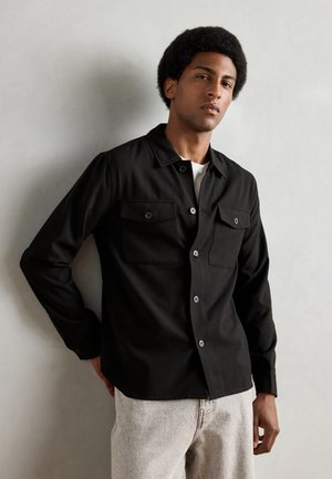 Black button-up shirt with two front pockets, featuring a collared neckline and long sleeves. Worn with light-colored, textured pants.