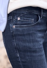 Gros plan sur la poche avant d'un jean en denim bleu foncé avec des coutures visibles et de petits rivets, porté avec un haut clair et une veste.
