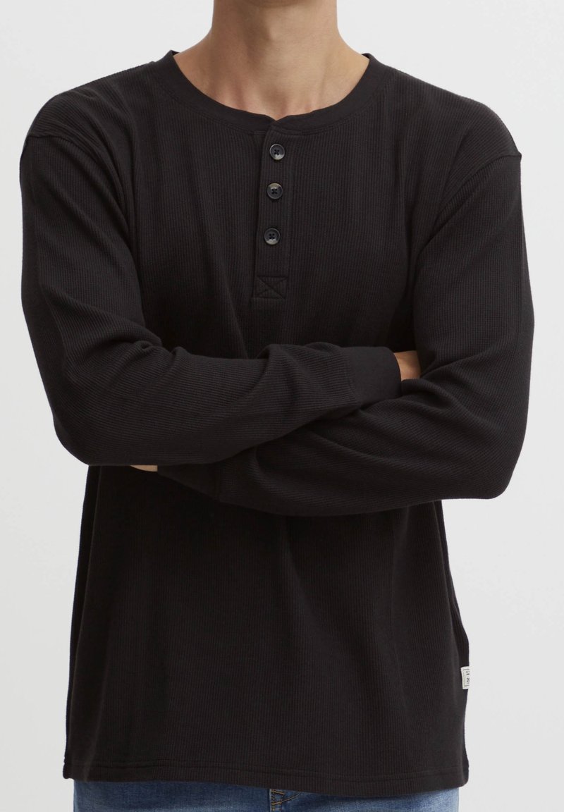 Personne portant un t-shirt Henley noir à manches longues côtelé avec trois boutons, bras croisés, visible des épaules jusqu'aux hanches.