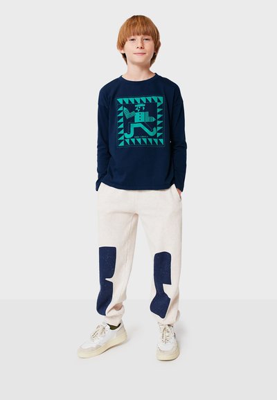 Marineblaues Langarmshirt mit geometrischem grünem Druck, kombiniert mit cremefarbenen Sweatpants mit marineblauen Patches. Weiße Sneakers vervollständigen das Outfit.