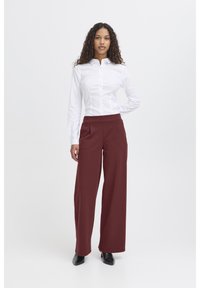 Witte blouse met knopen, gecombineerd met bordeauxrode wijde broek. De broek heeft plooien aan de voorkant, een gladde stof en een getailleerde pasvorm. Zwarte schoenen zichtbaar.