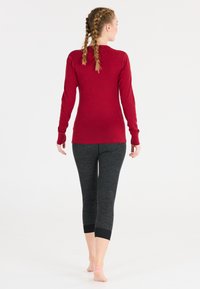 Kvinna med flätat hår som går barfota i en röd långärmad tröja och mörkgrå korta leggings mot en vit bakgrund.