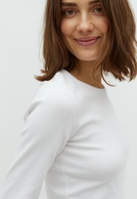 White, ribbed long-sleeve top with a crew neckline. The fabric appears soft and fitted, emphasizing the body's shape. Simple and minimal design.