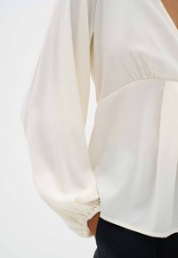 Blusa bianca con maniche lunghe e vestibilità rilassata. Presenta polsini arricciati e una texture liscia e leggera, ideale per essere indossata a strati.