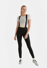 Leggings neri con bretelle, con una cintura gialla al neon. Il modello indossa una maglietta bianca aderente e scarpe da ginnastica. Tessuto elastico e liscio.