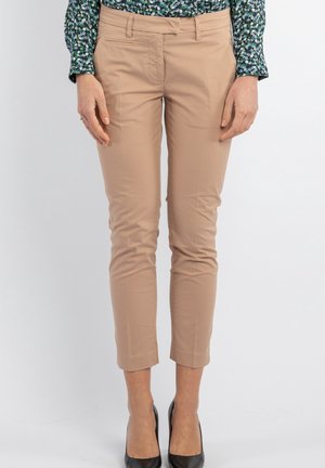 Kvinde iført beige slim fit cropped bukser, mønstret grøn og blå langærmet skjorte og sorte højhælede sko, står mod en ensfarvet baggrund.