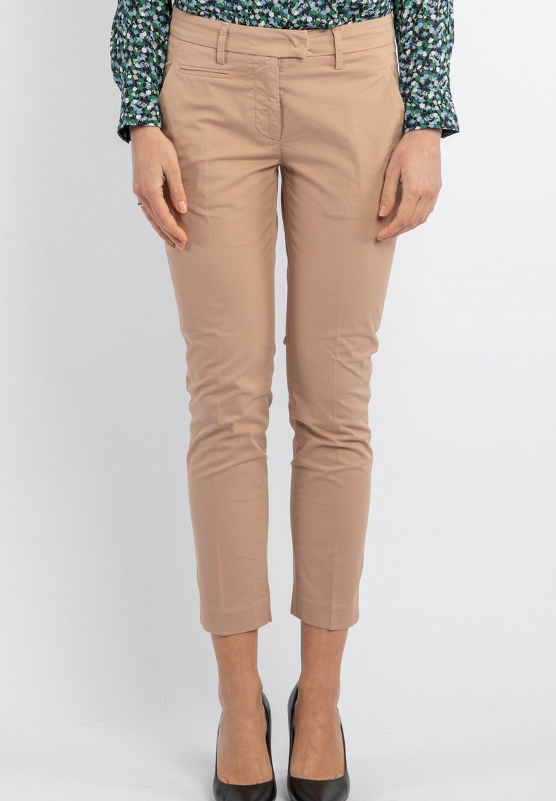 Femme portant un pantalon beige slim fit cropped, une chemise à manches longues à motifs verts et bleus, et des chaussures noires à talons hauts, debout devant un fonds uni.