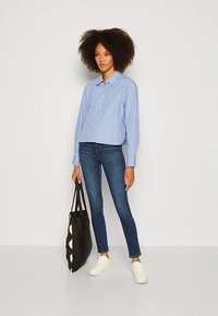 Modrá a bílá pruhovaná košile na knoflíky, doplněná přiléhavými tmavými džínami a bílými teniskami. Modelka drží černou tašku tote s vzorem.