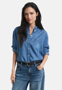 Denim button-up shirt in light blue, with long sleeves rolled up, a front pocket, and a relaxed fit. Paired with high-waisted blue jeans.