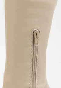 Bota de cuero beige hasta la rodilla con una cremallera lateral visible y un tirador texturizado, superficie lisa y costuras reforzadas en las uniones.