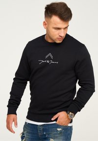 Black sweatshirt with round neck, long sleeves, and ribbed cuffs. Features a white logo and design on the chest, paired with distressed jeans.