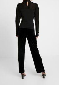 Schwarzes langärmeliges Oberteil mit gestreiften, transparenten Details und Schlüssellochausschnitt am Rücken, kombiniert mit schwarzen Samt-Hosen mit weitem Bein und Sandalen mit Absatz.