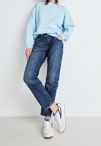Sudadera azul claro con un diseño gráfico, vaqueros azul oscuro con costuras blancas y zapatillas blancas con detalles en navy. Atuendo simple y casual.