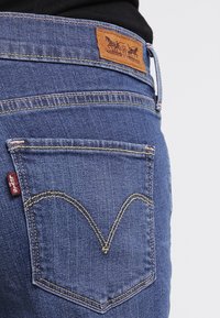 Bakficka på blå Levi's-jeans med brun läderlogotyp och röd tygetikett, båda slitna av person i svart topp.