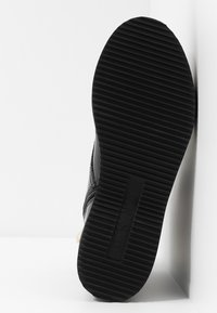 Semelle de chaussure noire avec des rainures horizontales et "DKNY" embossé au centre, posée à la verticale contre un fond blanc.