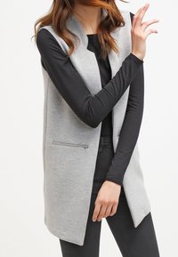 Jasnoszara kamizelka bez rękawów o kroju marynarki, z przednią kieszenią na zamek i gładką, teksturowaną tkaniną, zestawiona z czarną bluzką z długim rękawem.