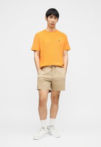 Moški stoji z rokami v žepih, oblečen v oranžno majico, bež kratke hlače, bele nogavice in bele superge na enobarvnem ozadju.