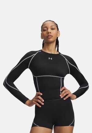 Atletische vrouw met gevlochten haar draagt een zwarte compressietop met lange mouwen en bijpassende shorts met grijze naadtoevoegingen, poserend met handen op de heupen.