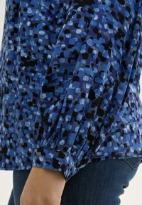 Langarmshirt in verschiedenen Blautönen mit einem gemusterten Design, das abgerundete Formen zeigt. Der Stoff ist leicht und hat geraffte Bündchen.