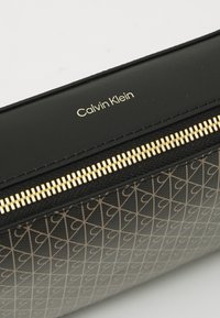 Juodas Calvin Klein piniginė su auksiniu užtrauktuku ir pasikartojančiu geometriniu raštu apatinėje dalyje.